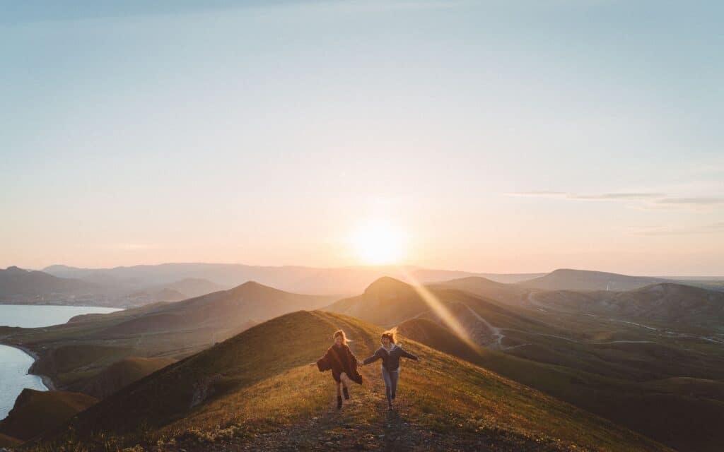 A Lua de Mel é o momento ideal para criar memórias. Casal de mulheres correndo de mãos dadas, com o pôr do sol ao fundo.  (Foto: Philipp Aleev/Unsplash)